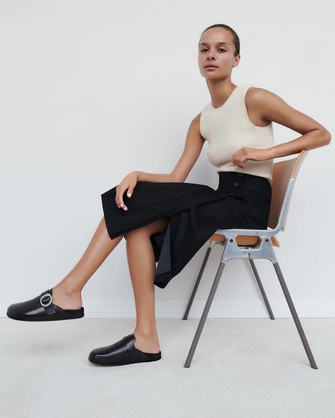 Elegant woman poses in a cream-colored top and black skirt, seated on a metal chair, wearing black leather mules in a bright indoor setting.