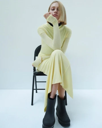 Model with blonde bob wearing a yellow top and skirt seated on a black chair against a white background
