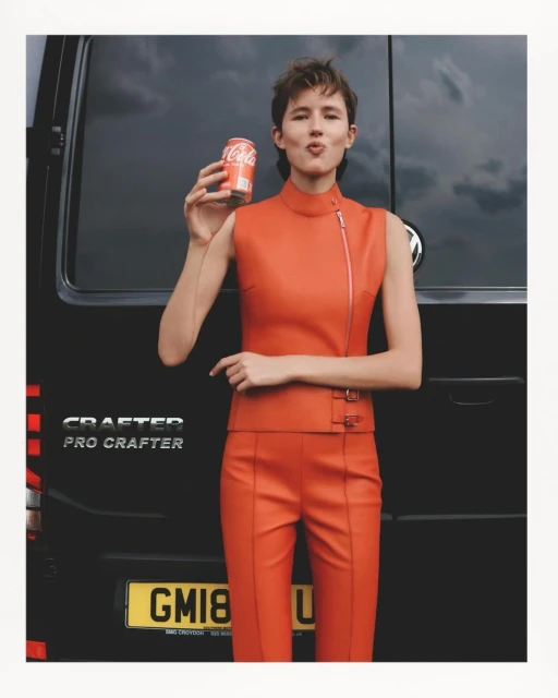 A woman in an orange pantsuit poses with a can of Coca-Cola in front of a black van, against an overcast sky.