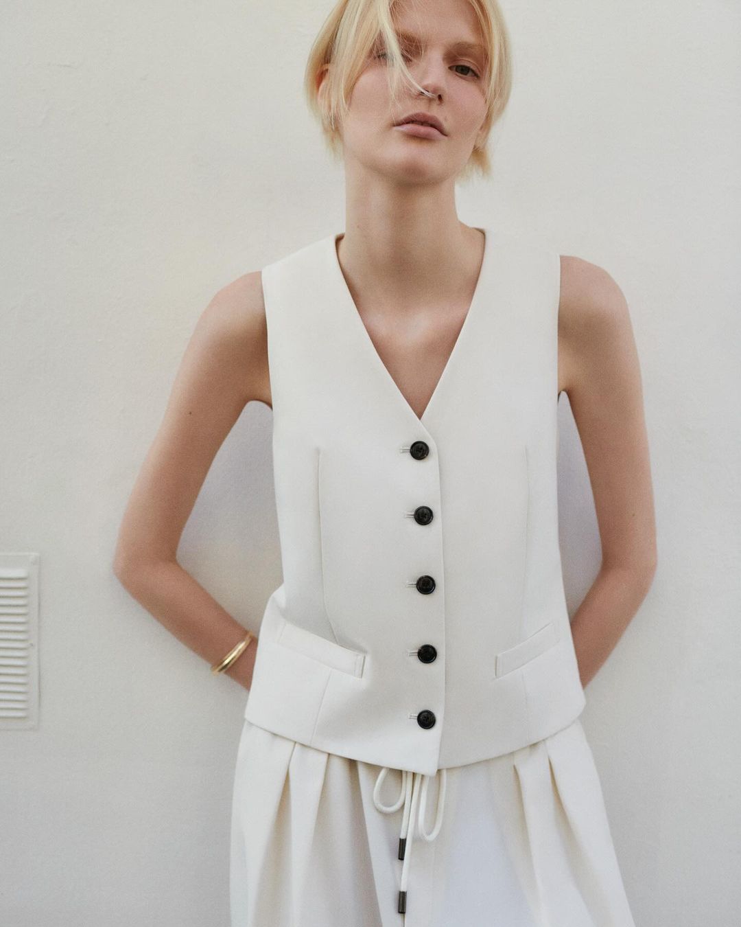 Fashion model in a sleeveless white suit vest and wide-leg pants against a bright neutral background, indoors, posing for a portrait.