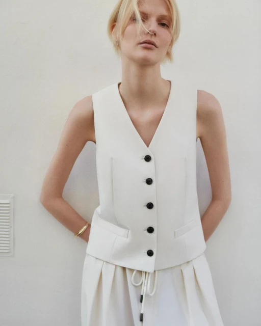 Fashion model in a sleeveless white suit vest and wide-leg pants against a bright neutral background, indoors, posing for a portrait.