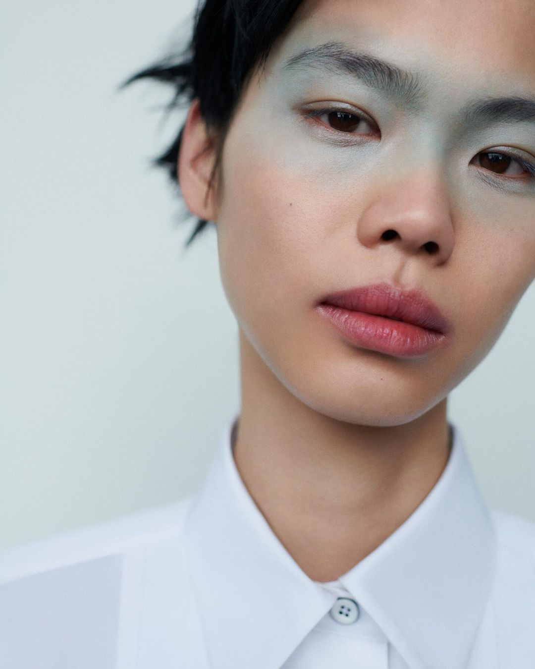 Close-up portrait of a young adult with light green makeup around the eyes, wearing a white shirt against a light background.