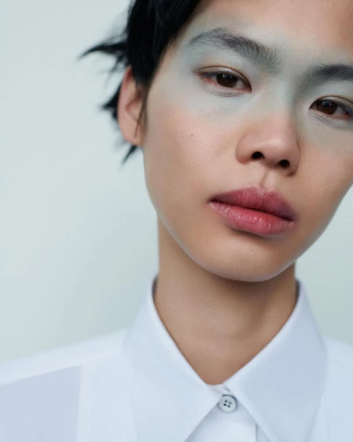 Close-up portrait of a young adult with light green makeup around the eyes, wearing a white shirt against a light background.