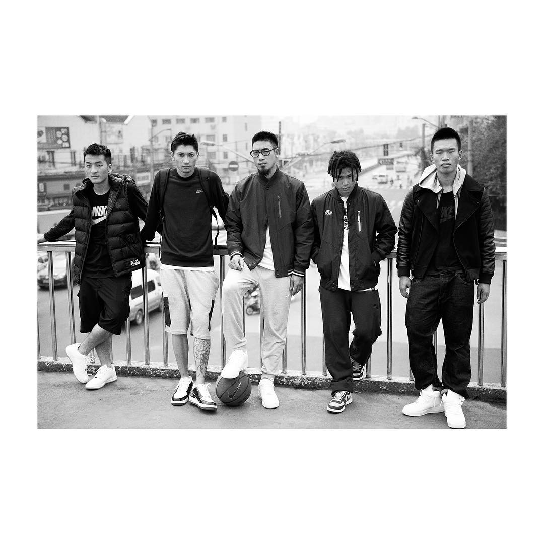Monochrome image of five young Asian men in jackets and athletic shoes standing outdoors with basketball, urban background.