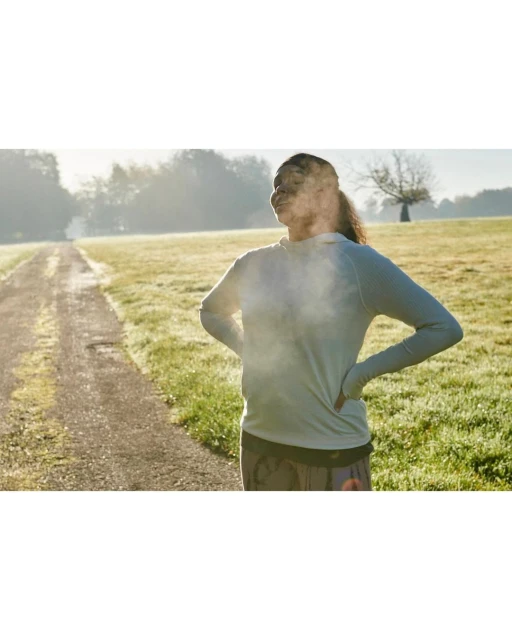 Woman in athletic wear stands outdoors on a path surrounded by a grassy field in bright sunlight, hands on hips taking deep breath.