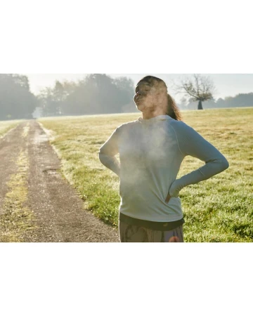 Woman in athletic wear stands outdoors on a path surrounded by a grassy field in bright sunlight, hands on hips taking deep breath.