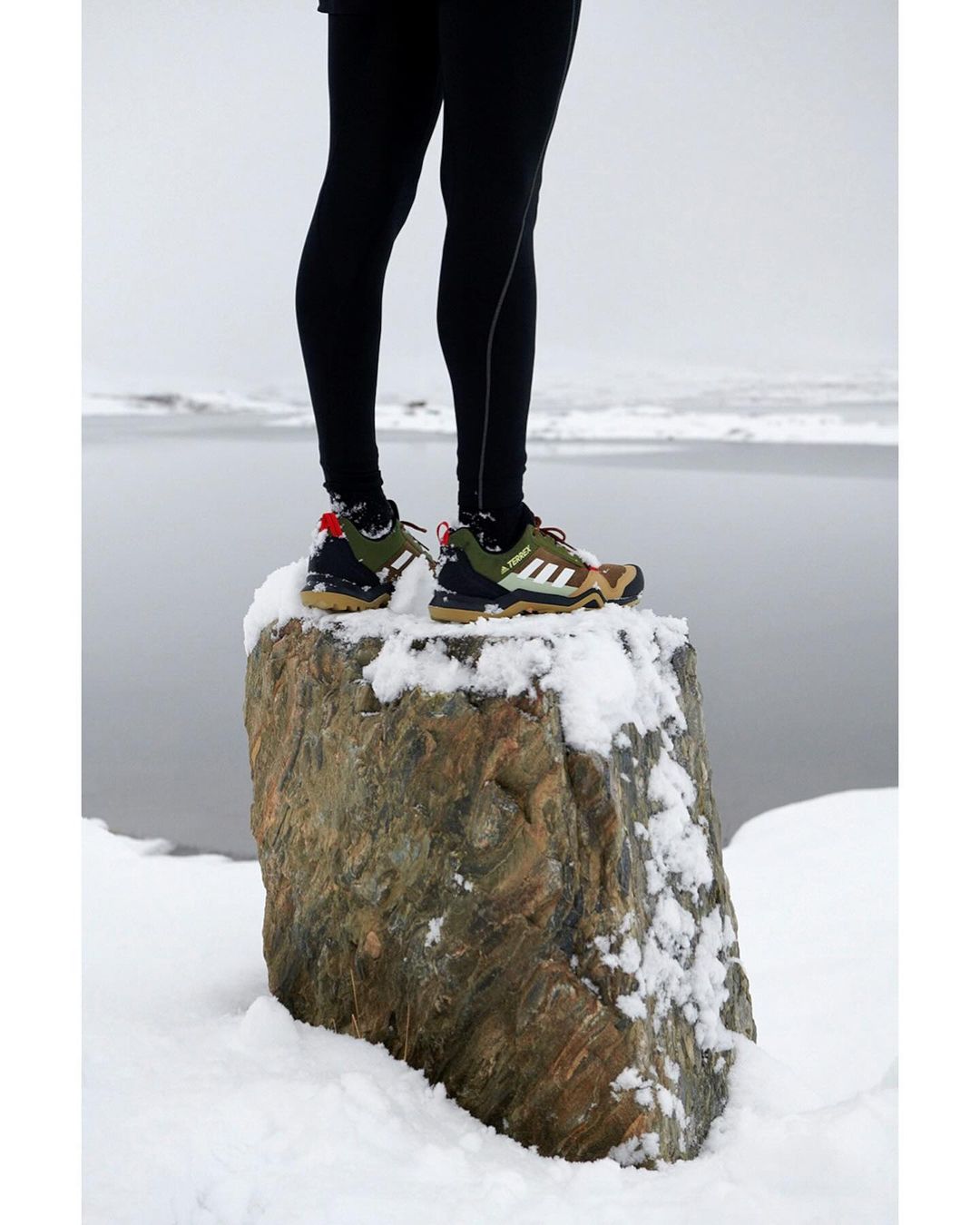 Legs in black leggings and Adidas trail running shoes, standing on a snow-covered rock with a lake and overcast sky in the background.