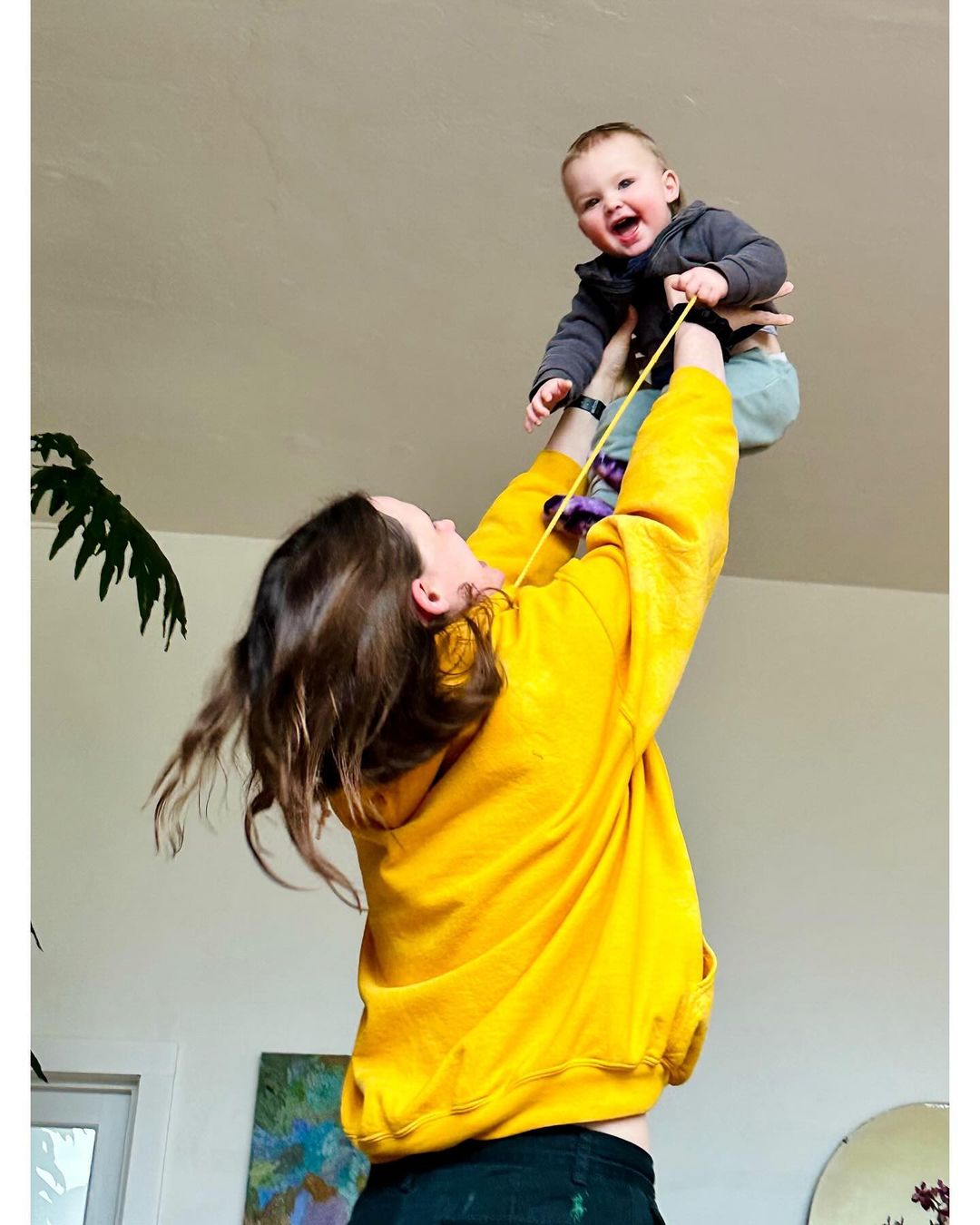 A mother in a yellow sweatshirt lifts her smiling baby into the air in a brightly lit indoor space, with a plant and painting visible.