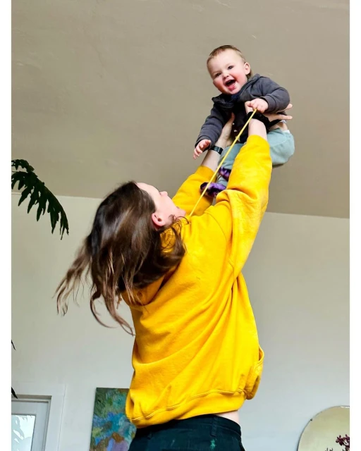 A mother in a yellow sweatshirt lifts her smiling baby into the air in a brightly lit indoor space, with a plant and painting visible.