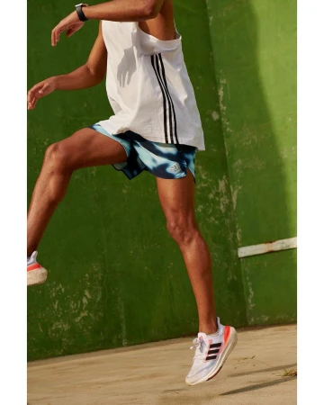 Man wearing a white tank top and blue patterned running shorts mid-air against a weathered green wall.