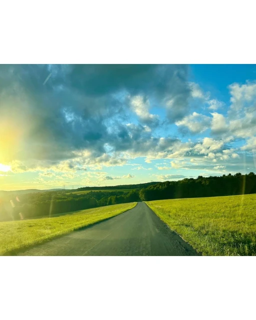 Scenic view of a black asphalt road cutting through green grassy fields and leading to a distant treeline under a partly cloudy sky.