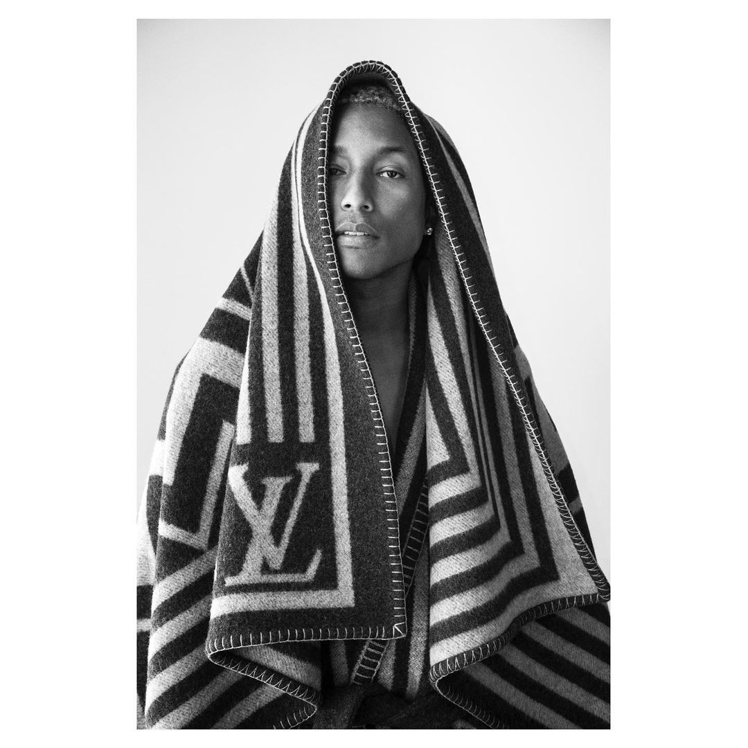 Monochrome studio portrait of an adult wearing a striped Louis Vuitton shawl wrapped around the head and body against a white backdrop.