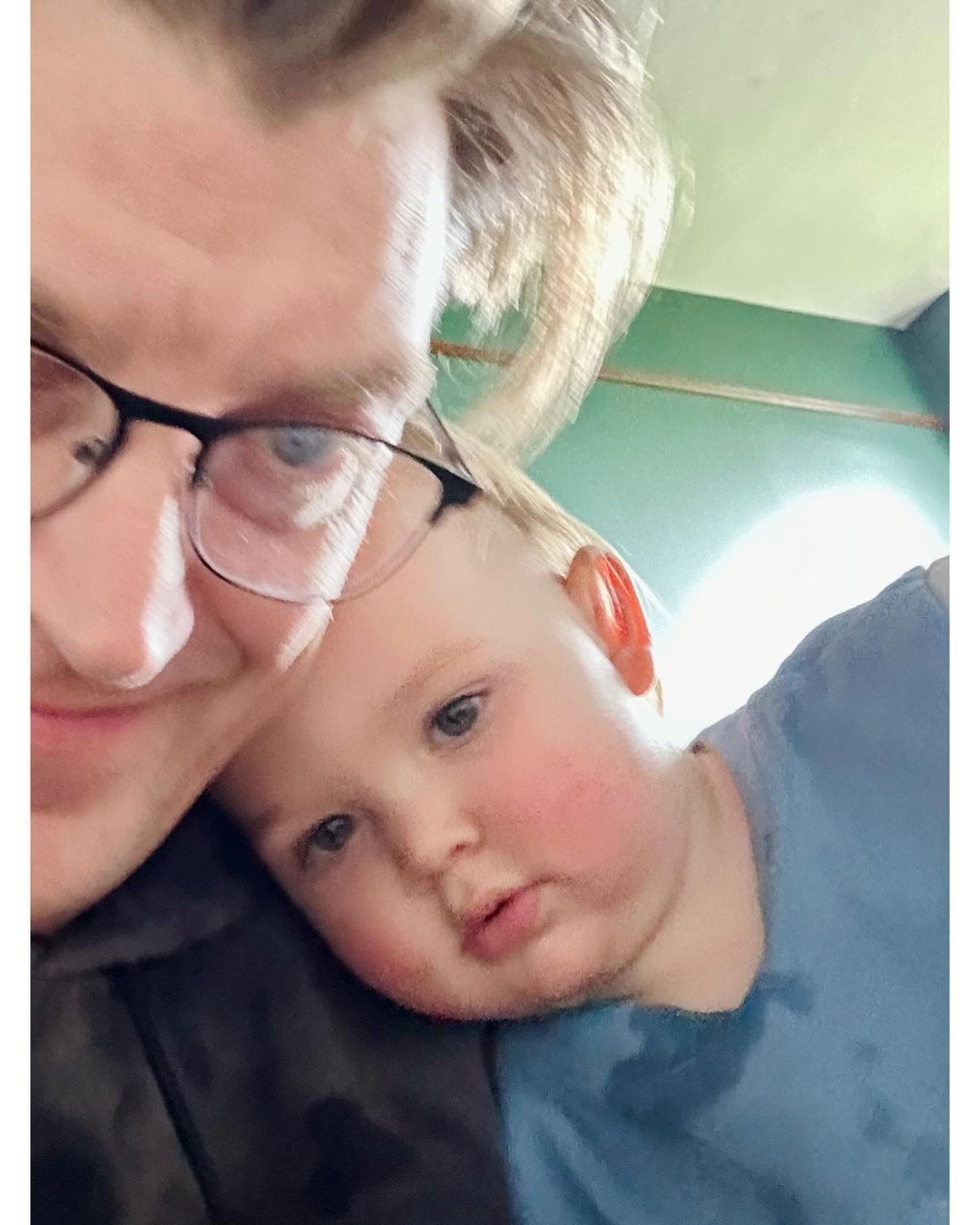 Close-up of a fair-skinned adult male wearing glasses and a fair-skinned child with blond hair in a blue shirt indoors.