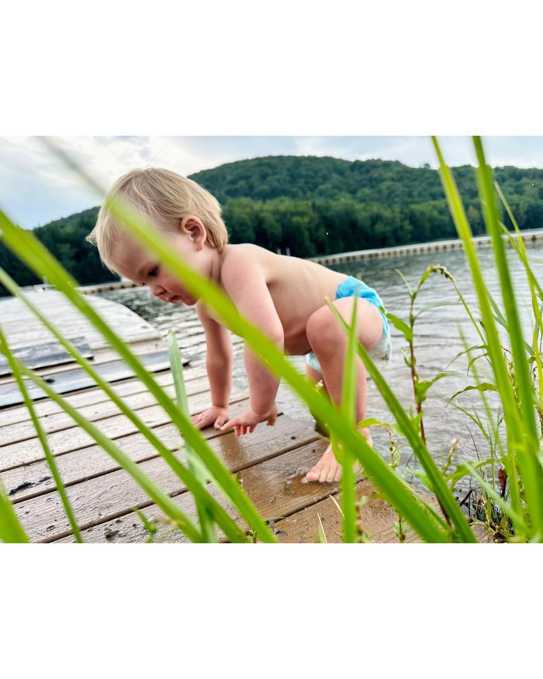 Toddler explores on a wooden dock by a lake with grassy foreground.  Green hills are visible in the background under a cloudy sky.