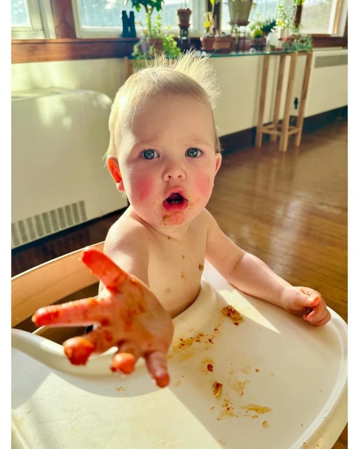 Close-up of a baby in a high chair covered in spaghetti sauce reaching out with a messy hand in a well-lit indoor setting.
