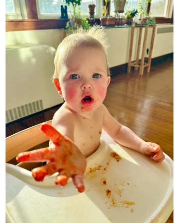 Close-up of a baby in a high chair covered in spaghetti sauce reaching out with a messy hand in a well-lit indoor setting.