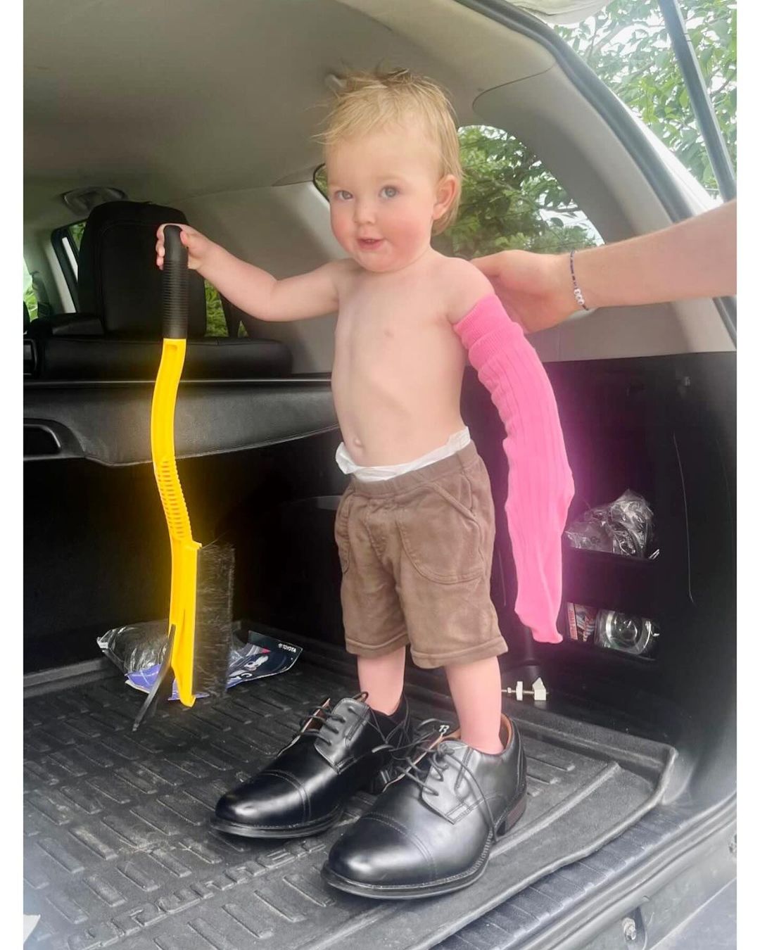 Toddler stands in a car trunk wearing oversized dress shoes and holding a yellow brush, while someone places a pink sleeve on their arm.