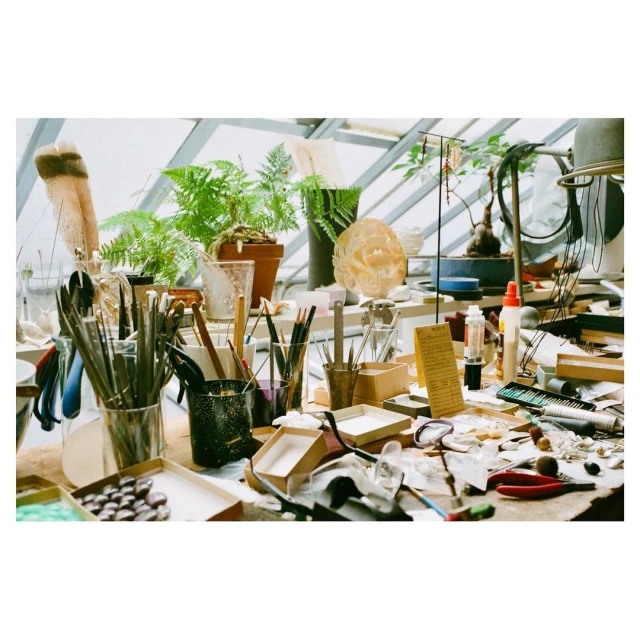 Close-up of a cluttered worktable with crafting tools, beads, boxes, paint, and plants in a sunlit workshop, showcasing an artistic environment.