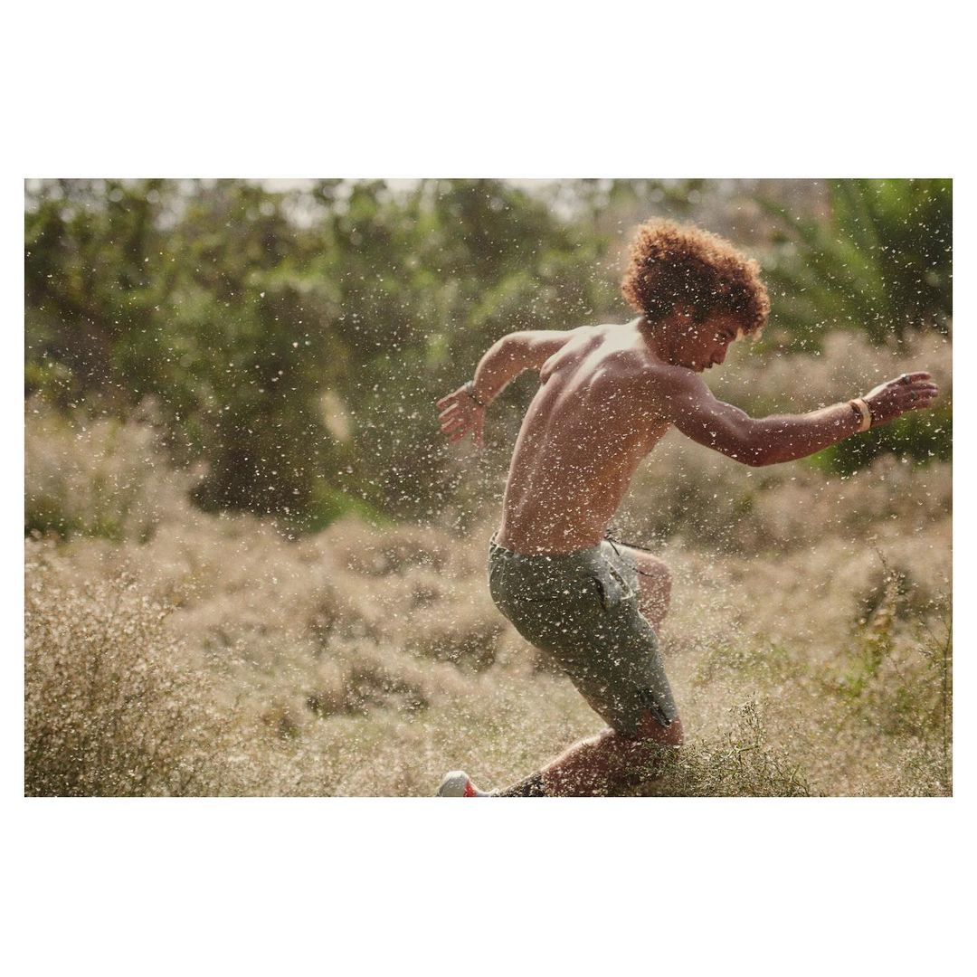 Athletic man with curly red hair running through a field of tall, dry grass with a blurred background of green trees.