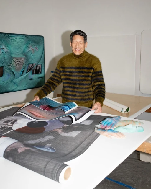 Man holding large printed artwork indoors with digital art display on the wall. Smiling Asian male artist in creative studio space.