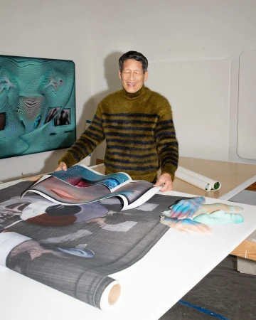 Man holding large printed artwork indoors with digital art display on the wall. Smiling Asian male artist in creative studio space.