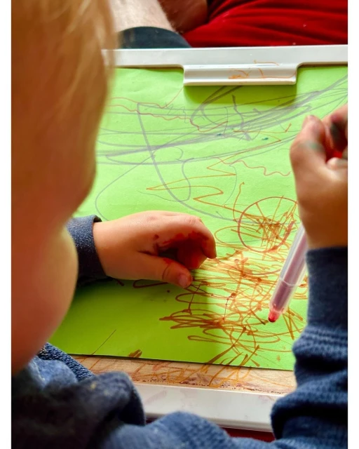 Toddler concentrates on drawing colorful scribbles on bright green paper with a red marker, viewed from behind in an indoor setting.