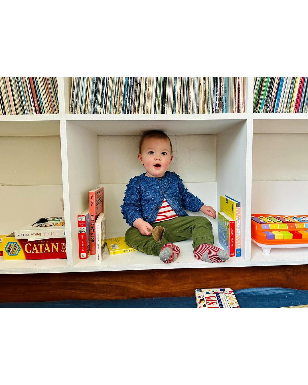 Infant sits in a white bookshelf with books and toys, wearing a navy blue sweater and green pants, looking directly at the camera.