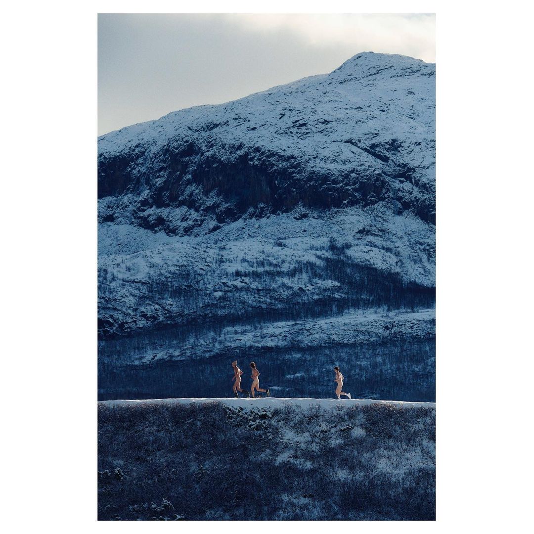 Three adult figures running in a snow landscape against a backdrop of a mountain covered in snow under an overcast sky.