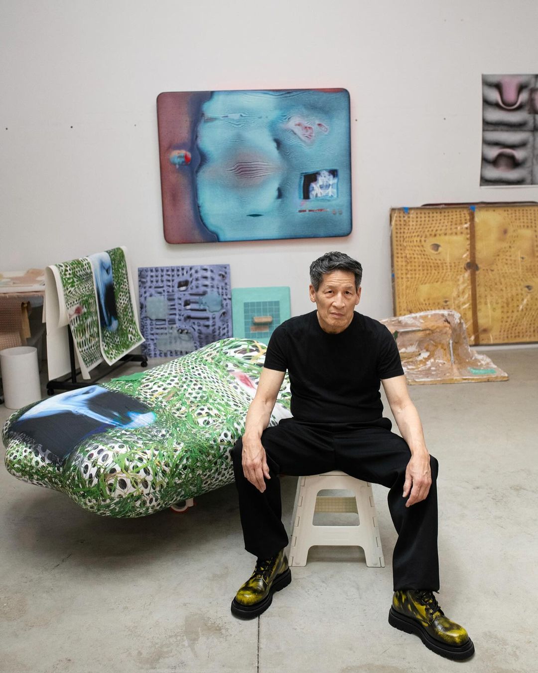 Artist in studio poses with his art. Man sits on stool surrounded by paintings and art objects in brightly lit creative space.