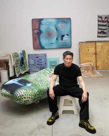 Artist in studio poses with his art. Man sits on stool surrounded by paintings and art objects in brightly lit creative space.