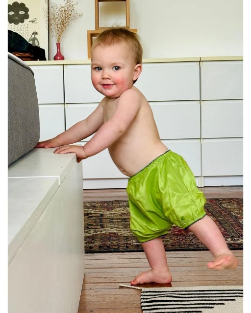 Infant with fair skin and blonde hair wearing lime green bloomers stands by white furniture inside a home with a brown and white rug.