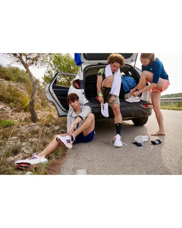 Three young adults lace up Adidas sneakers after a run, gathered around the open trunk of a white SUV on a rural road.