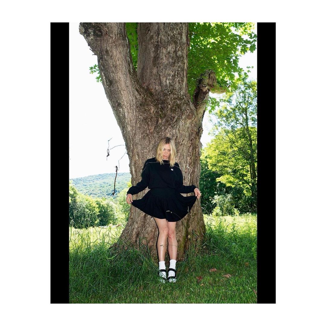 Woman in a black dress standing against a large tree trunk in a grassy field with trees under bright sunlight