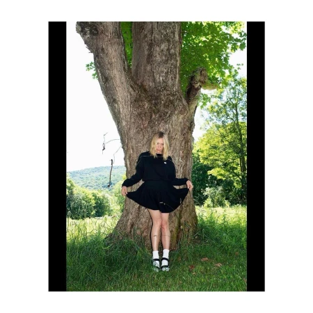 Woman in a black dress standing against a large tree trunk in a grassy field with trees under bright sunlight