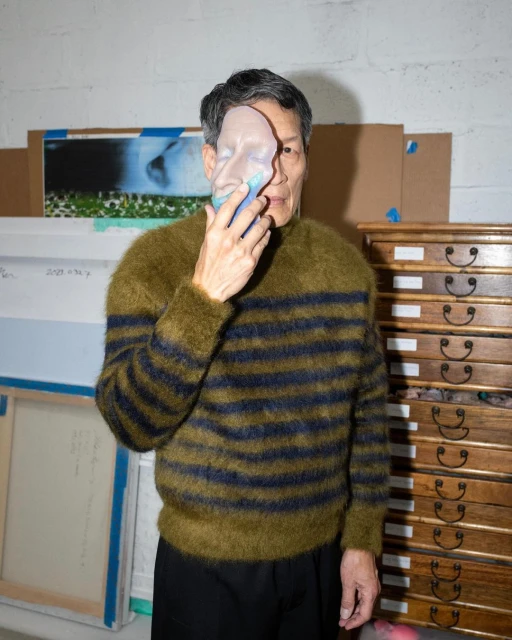 Asian man poses indoors with striped sweater and mask in art studio filled with art supplies and wooden drawers against a white brick wall.