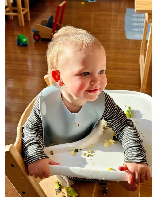 Close-up of baby in high chair wearing bib and striped long sleeve shirt, eating with food visible on tray and wooden floor.