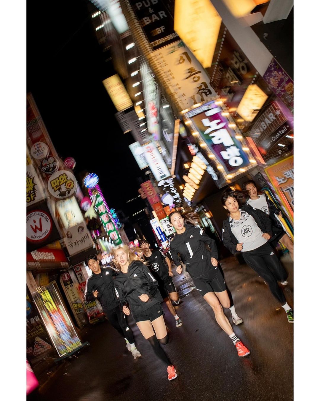 Group of people running in urban city street at night. Multiple adults are wearing sporty athletic clothing with city lights. 