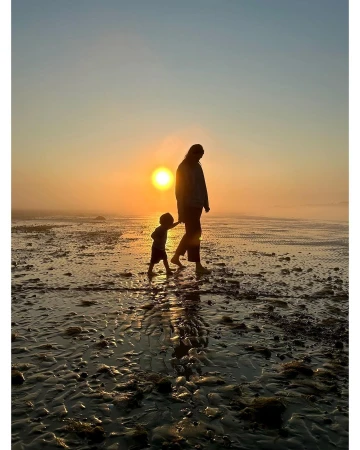 Silhouette of an adult and a young child walking together on a wet sandy beach during a bright sunset.