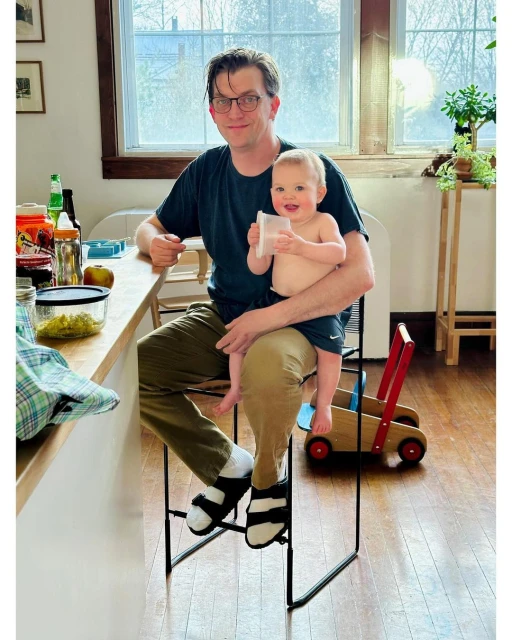 A man wearing glasses and holding a baby in his lap, both smiling, with a child's walker and indoor kitchen setting.