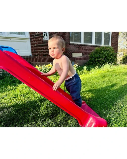 A toddler climbs a red plastic slide set on a green lawn in front of a brick house, enjoying outdoor playtime in bright daylight.