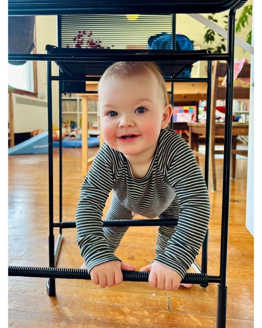 Adorable baby with a happy expression crawls under a black metal table, creating a playful and engaging moment in a brightly lit room.