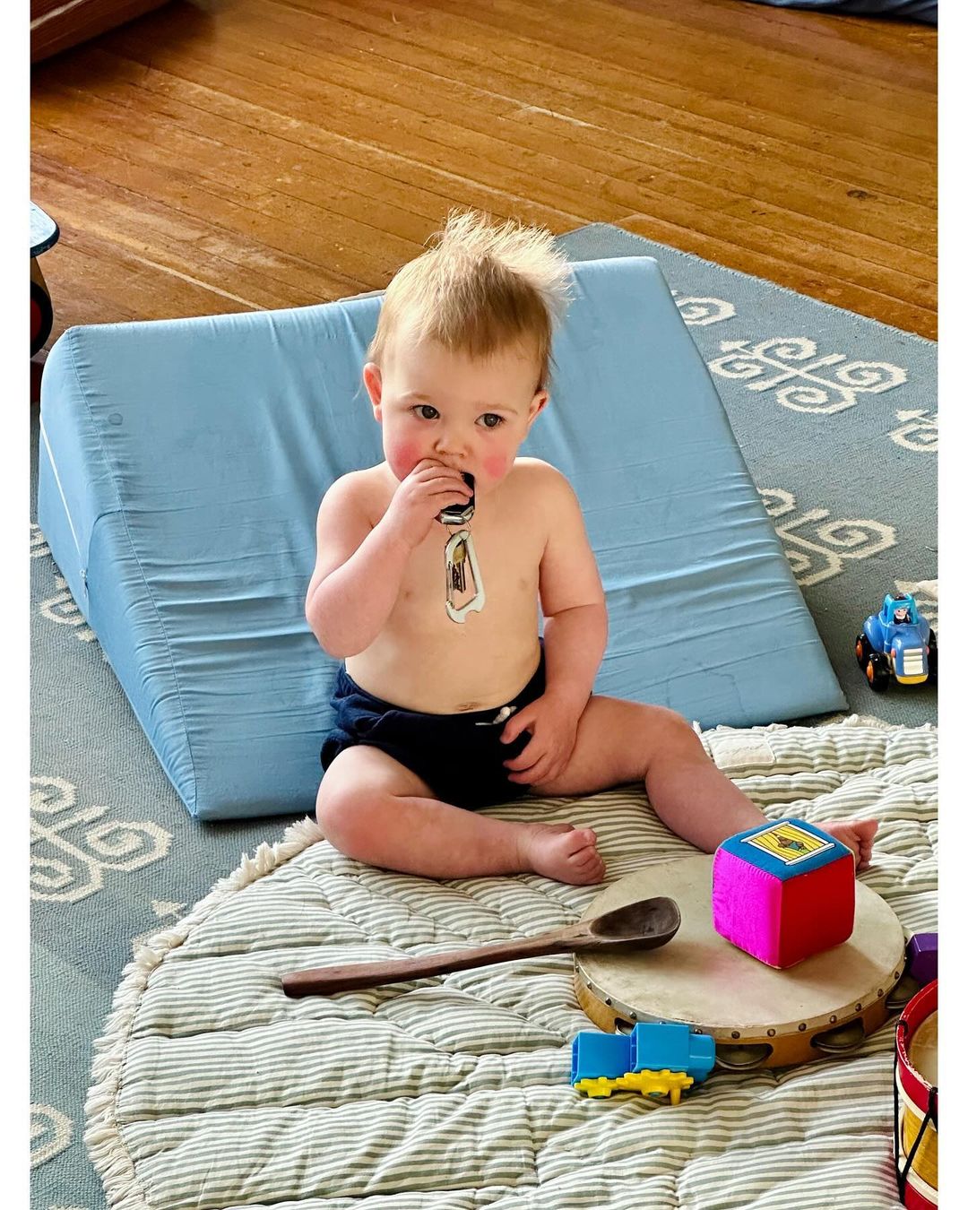 Baby sits surrounded by toys on a patterned rug, with a blue wedge pillow in the background, looking at the camera.