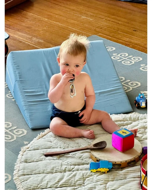 Baby sits surrounded by toys on a patterned rug, with a blue wedge pillow in the background, looking at the camera.