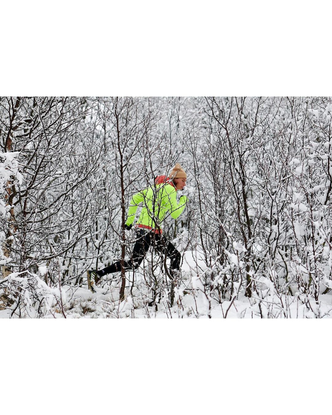 A person in athletic wear runs through a snow-covered forest, wearing a neon yellow jacket and hat, surrounded by trees and snow.