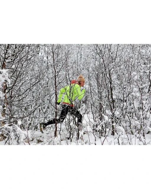 A person in athletic wear runs through a snow-covered forest, wearing a neon yellow jacket and hat, surrounded by trees and snow.