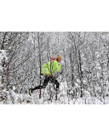 A person in athletic wear runs through a snow-covered forest, wearing a neon yellow jacket and hat, surrounded by trees and snow.