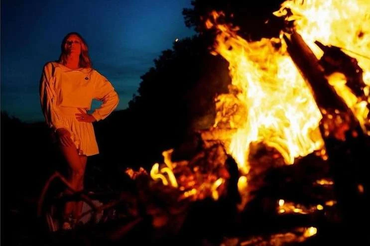 Woman posing near bonfire at night. Campfire flames light her white outfit against the dark blue sky. Woman stands confidently.