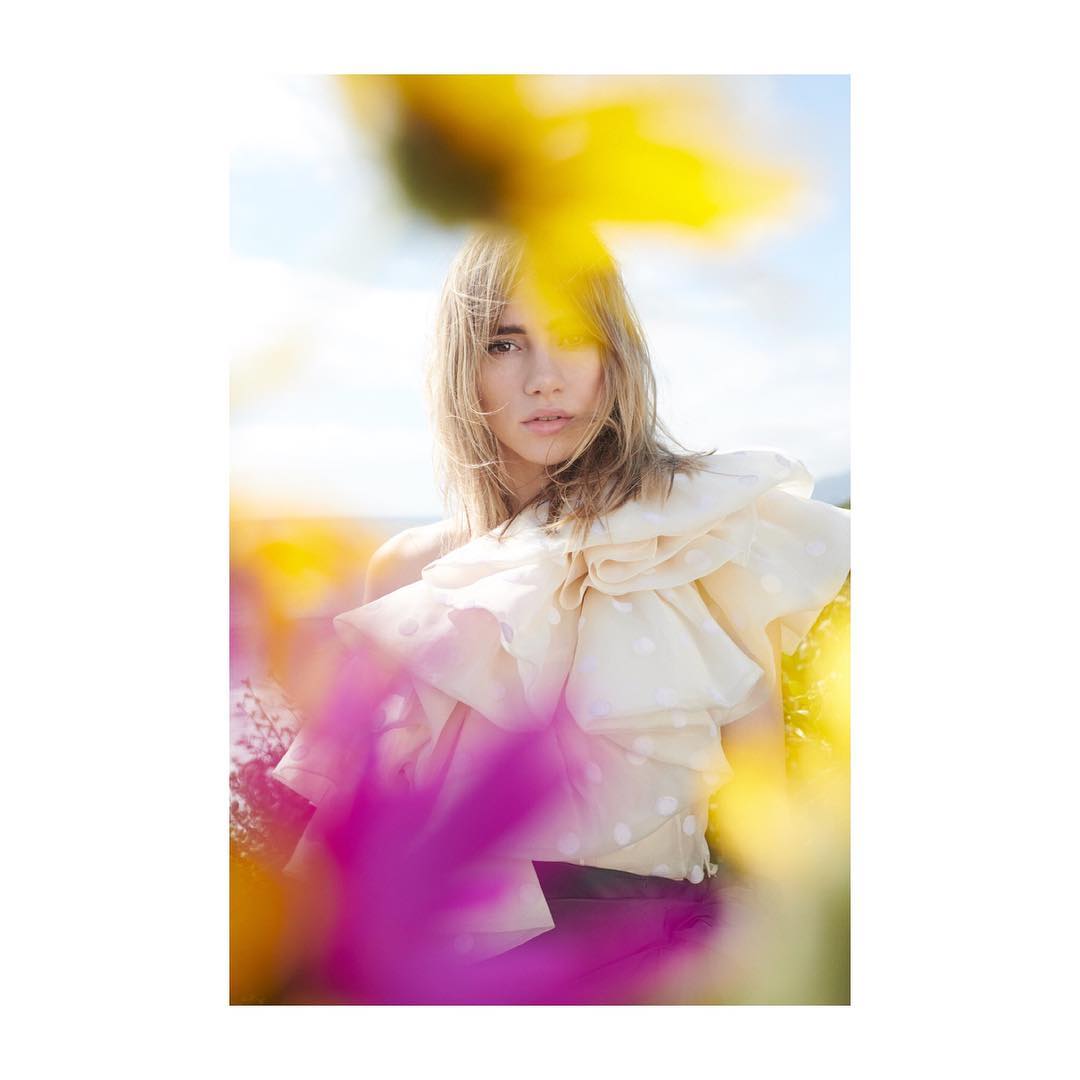 Portrait of a woman wearing a large ruffled blouse, standing in a field of colorful wildflowers under a bright, sunny sky.