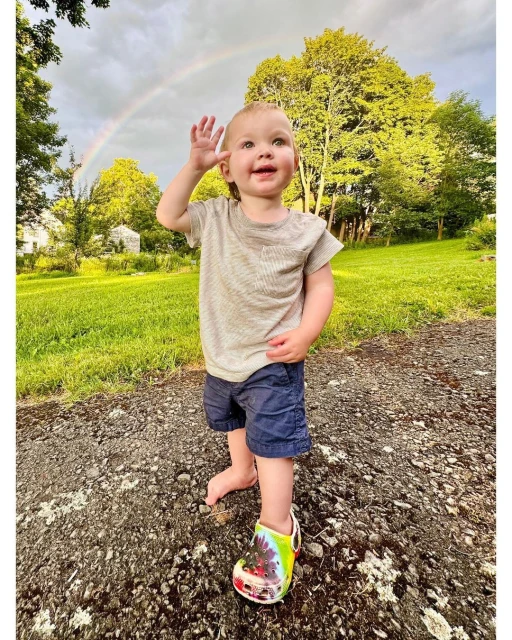 Toddler waves outdoors with a rainbow in the background. The child wears shorts and a t-shirt while standing on a gravel path.