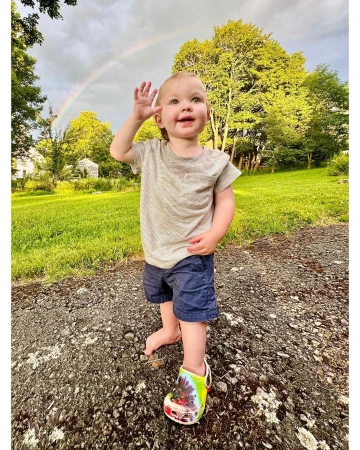 Toddler waves outdoors with a rainbow in the background. The child wears shorts and a t-shirt while standing on a gravel path.